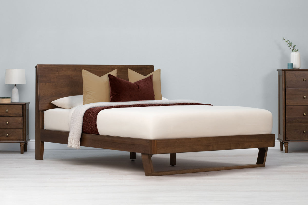 Mayfair solid walnut double bed frame with floating sled leg, angled view with cream bedding, cushions and matching walnut furniture