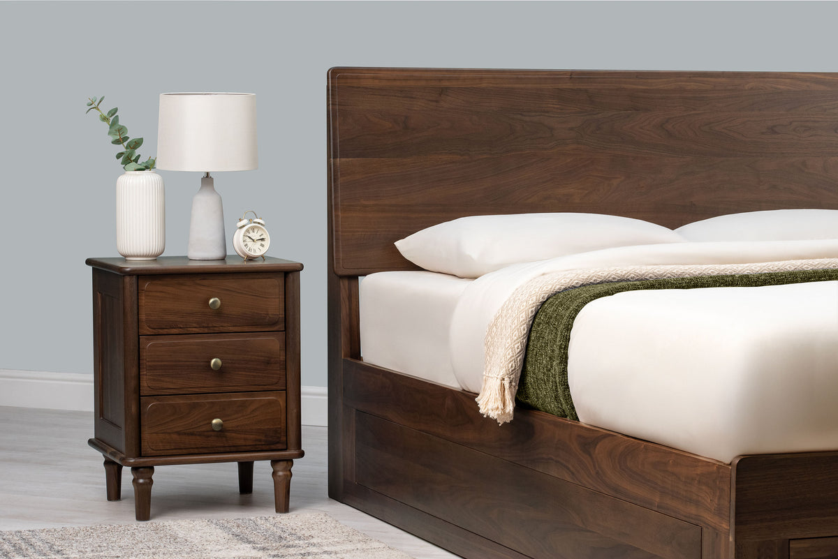 Mayfair solid walnut double ottoman bed headboard with plain white pillows, beside a matching three-drawer bedside table