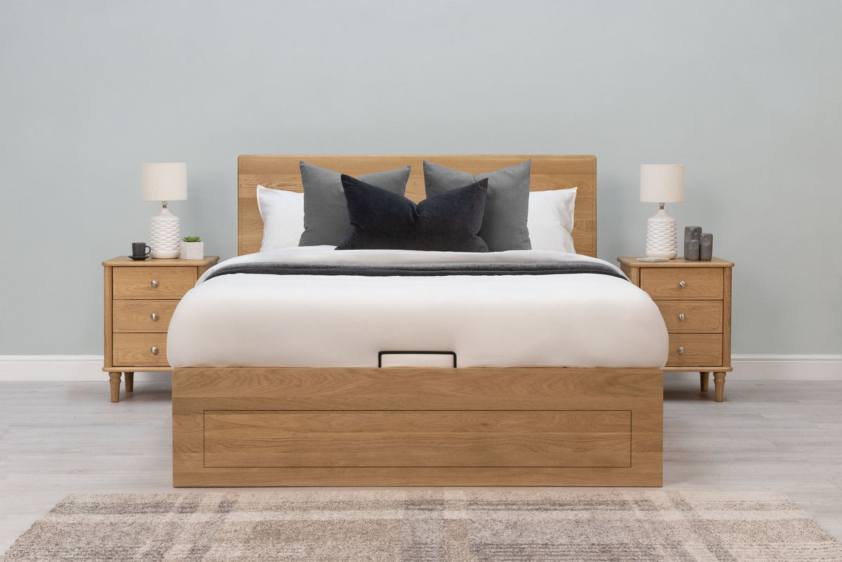 Mayfair Solid Natural Oak Ottoman Storage Bed Frame 4ft6 Double, front view dressed with white bedding and grey cushions beside oak bedside tables