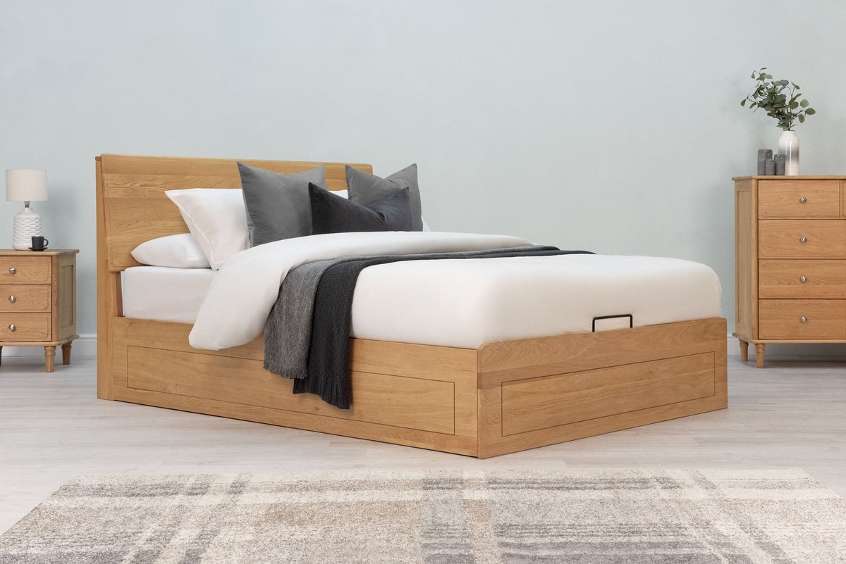 Mayfair Solid Natural Oak Ottoman Storage Bed Frame 4ft6 Double, side angle view dressed with white bedding, grey cushions and a grey throw beside an oak bedside table and chest of drawers