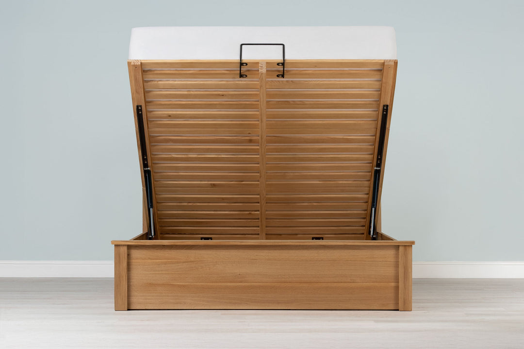 Wilmore solid natural oak double ottoman bed frame with a bare mattress and the base raised, front view from the foot of the bed