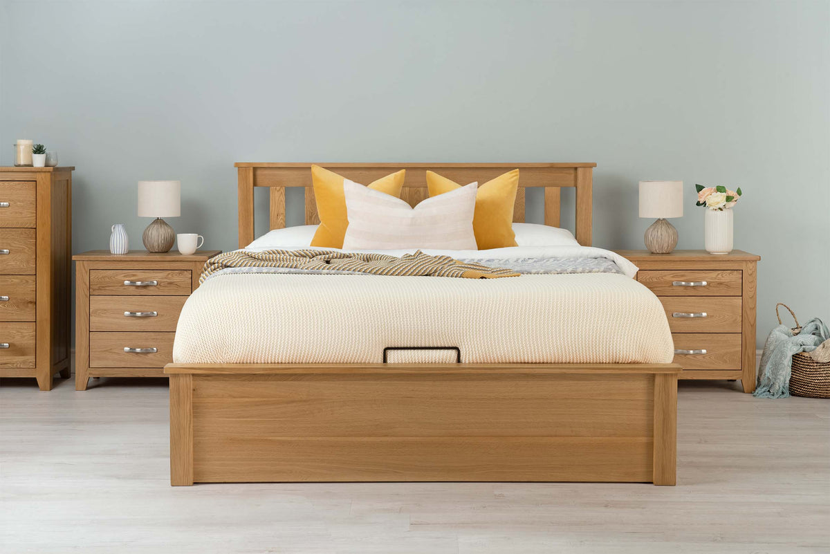 Wilmore solid natural oak king size ottoman bed frame dressed with cream bedding and mustard cushions, front view with matching bedside tables