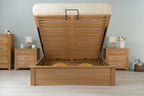 Wilmore solid natural oak king size ottoman bed frame with the mattress base raised, front view showing the under-bed storage in a dressed bedroom