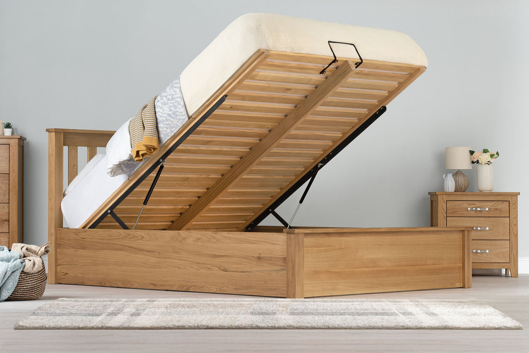 Wilmore solid natural oak king size ottoman bed frame with the mattress base raised on gas-lift struts, side view in a dressed bedroom