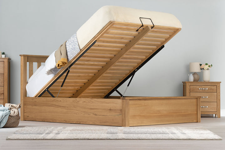 Wilmore solid natural oak super king ottoman bed frame with the mattress base raised on gas-lift struts, side view in a dressed bedroom